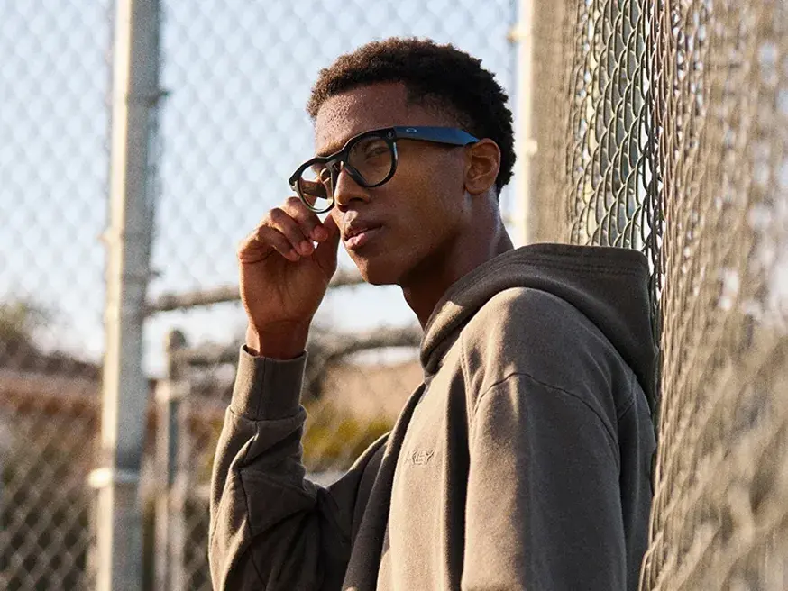 A person wearing a dark hoodie and smart sunglasses stands next to a wire fence outdoors, holding the glasses while sunlight filters through the background.
